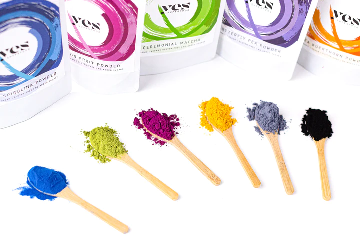 A Yes Superfood szivárvány színű porai fakanalakra adagolva: kék spirulina, sárkánygyümölcs, matcha, pillangóborsó, kurkumin, fekete répa és aktív szén – mindegyik természetes ételszínezék és tápanyagforrás egyben.