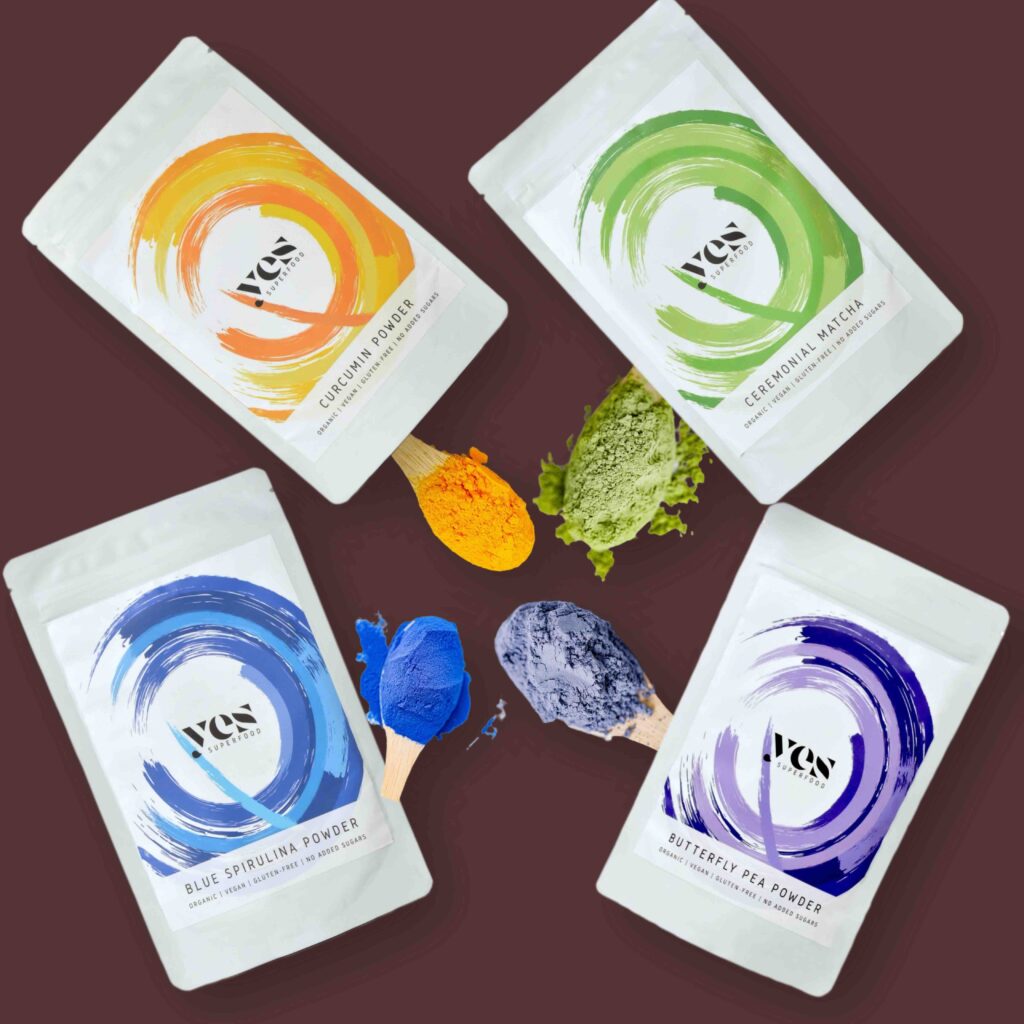 Yes Superfood Curcumin, Matcha, Blue Spirulina és Butterfly Pea porok kreatív, színes kompozícióban fa kanalakkal.